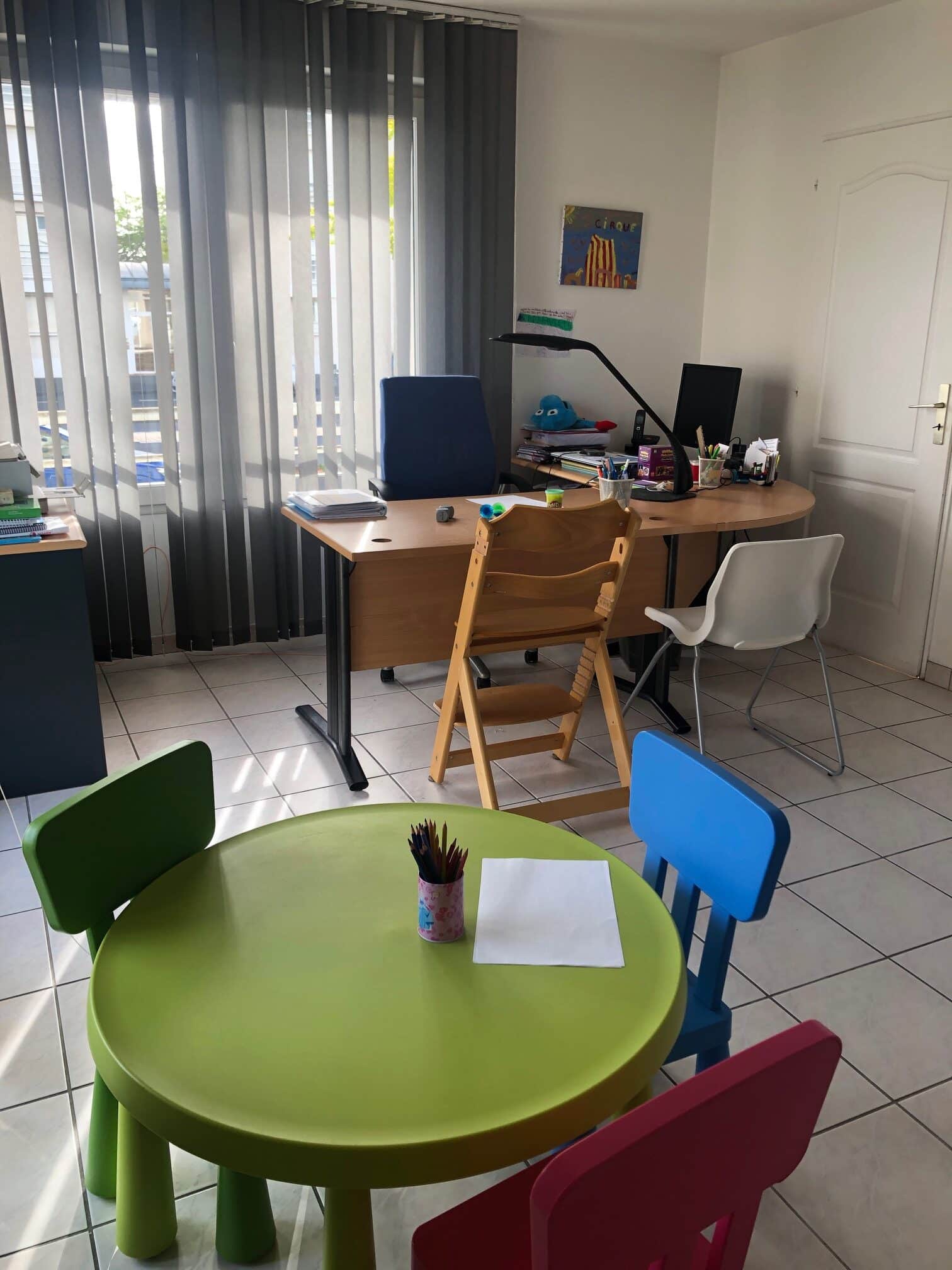 Photo du cabinet de Sophie Rossignol graphopédagogue à Caen. Un grand bureau avec un fauteuil pour la graphopédagogue. L'élève s'installe en face sur une chaise type Trip Trap selon sa taille. Une autre chaise est à disposition à côté pour l'éventuel parent qui accompagne.